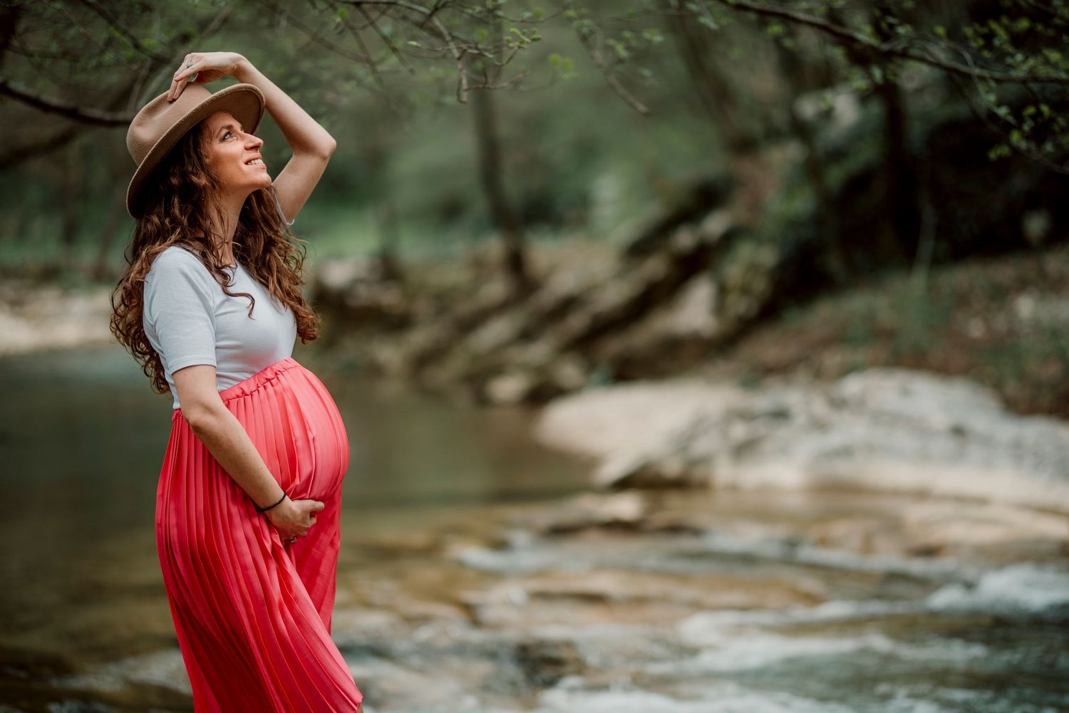 Pregnant woman in red skirt and cowboy hat poses gracefully by a rocky creek in a lush forest setting.