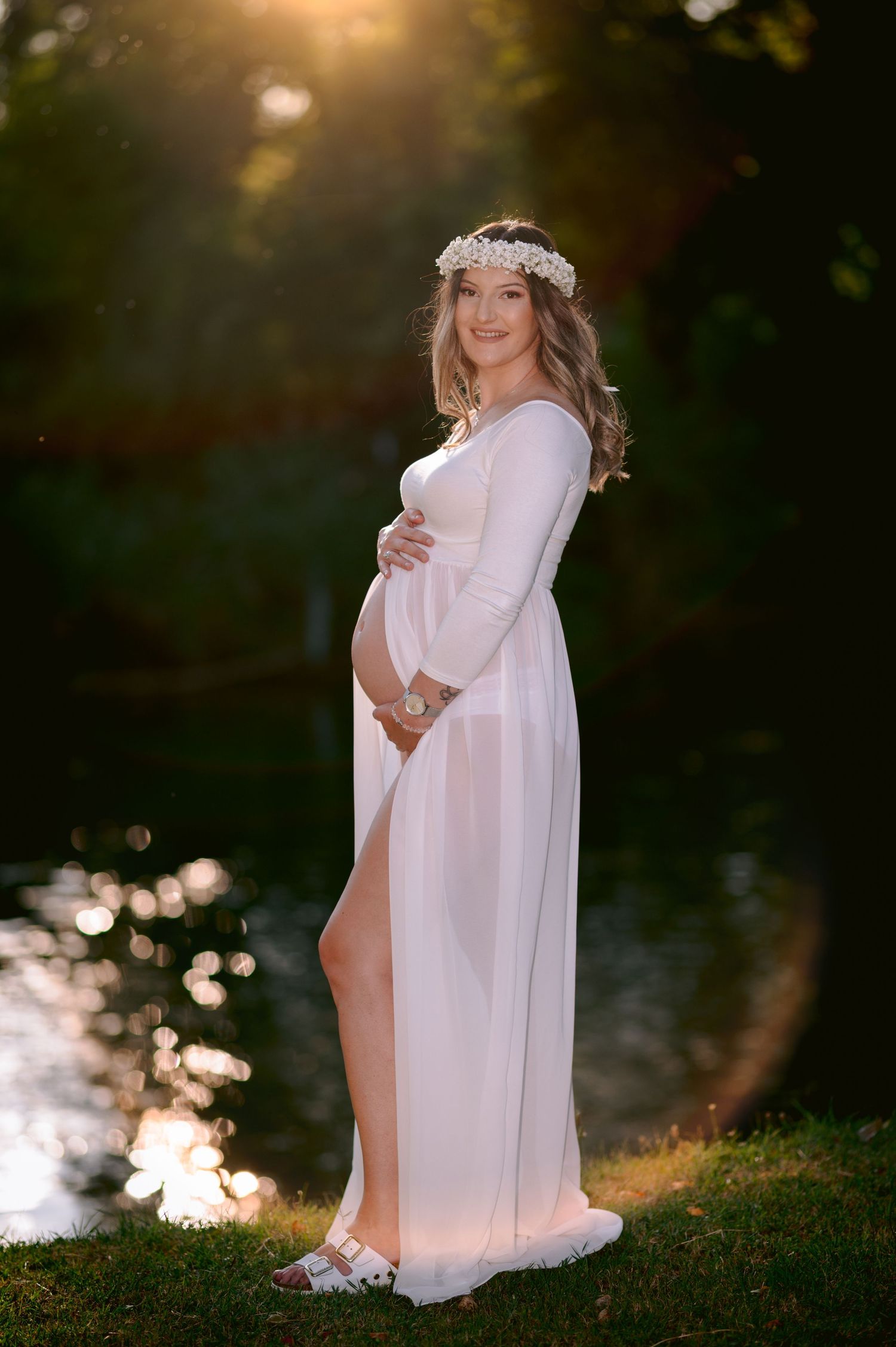 Pregnant woman in white gown and floral crown poses outdoors at golden hour by a sunlit pond.