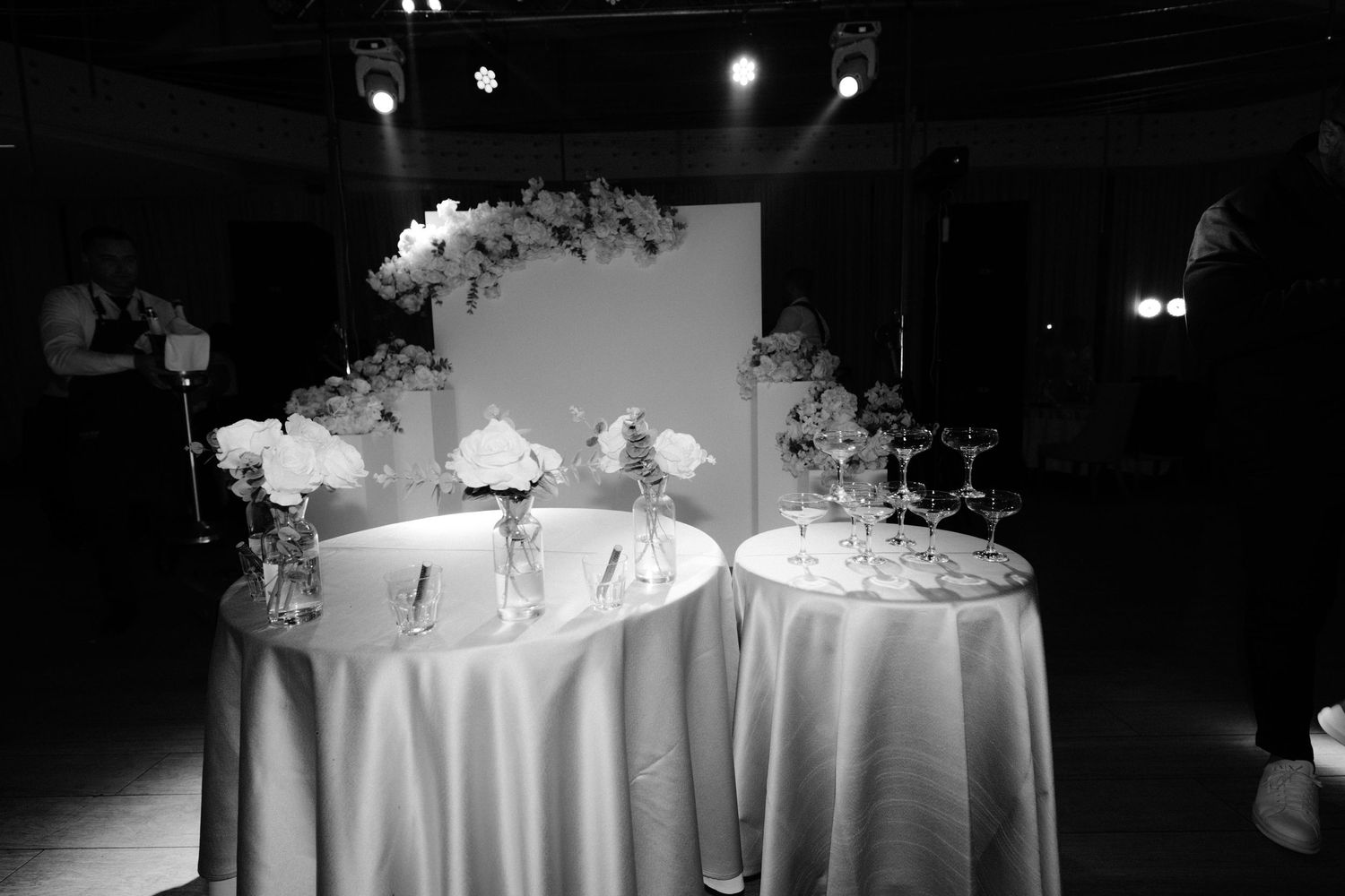 Elegant wedding reception tables with white linens and floral centerpieces illuminated by soft lighting in a dark venue.