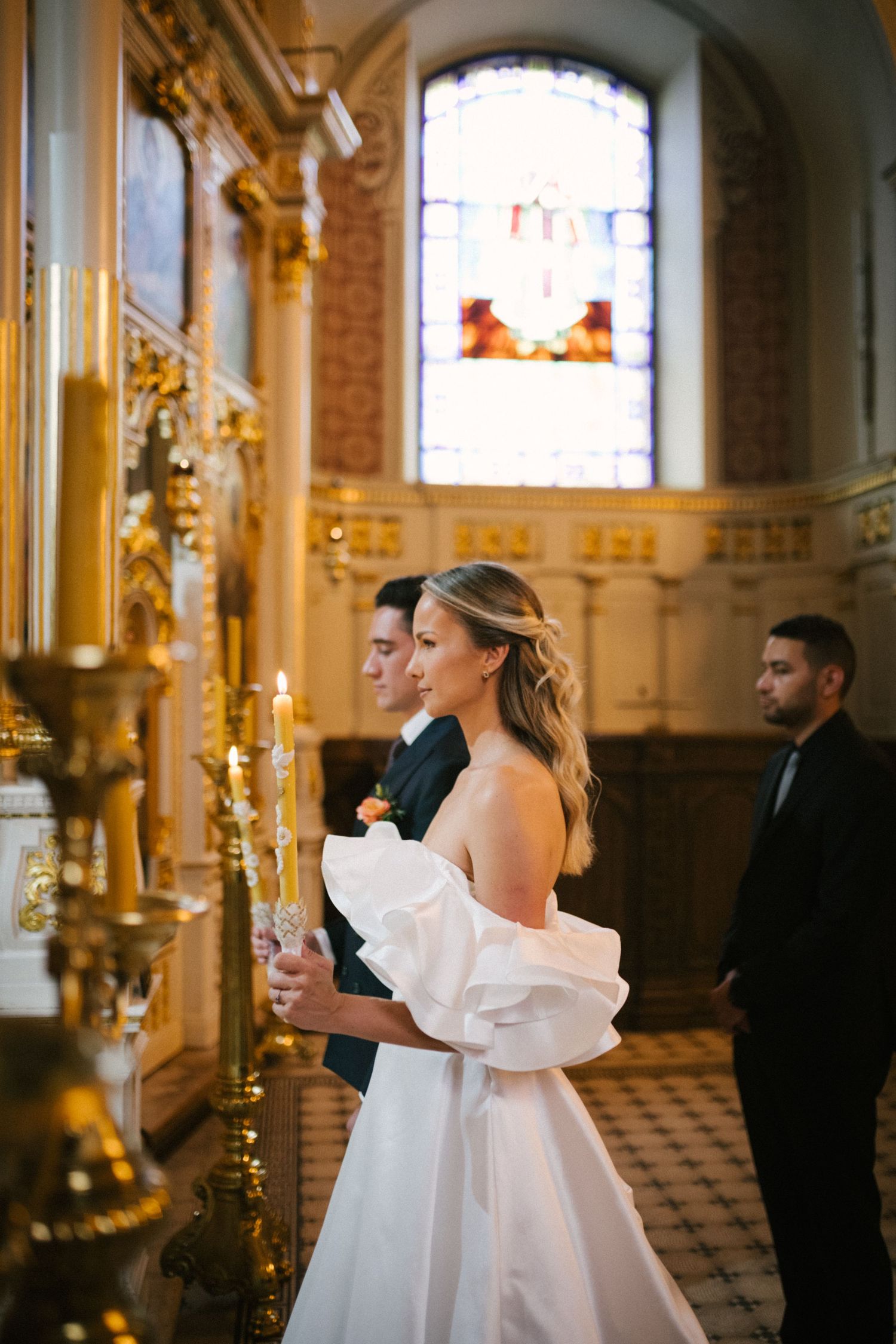 A couple stands in an ornate church with stained glass windows and golden candlesticks during their wedding ceremony.