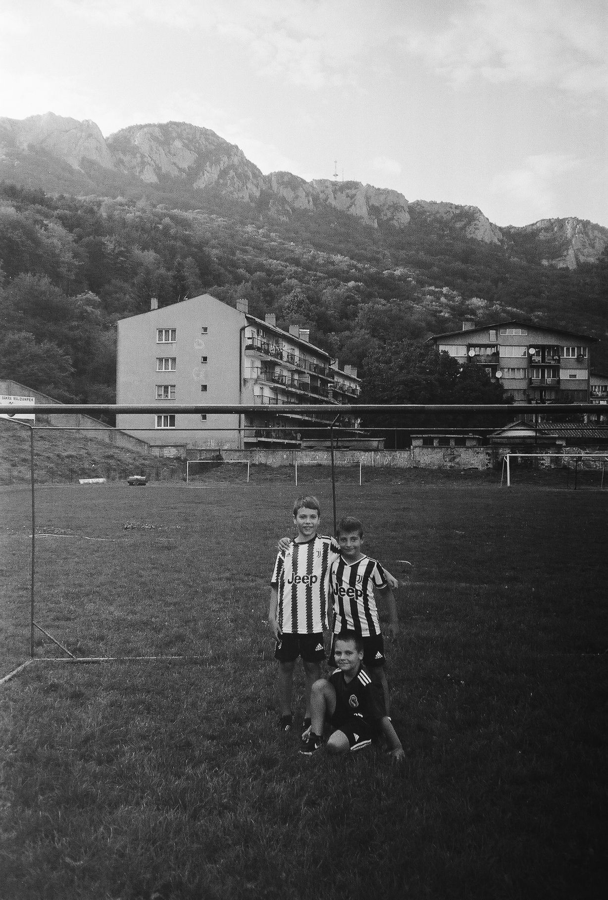 Black and white sequence of soccer players in striped jerseys practicing on a field with buildings and mountains in background.