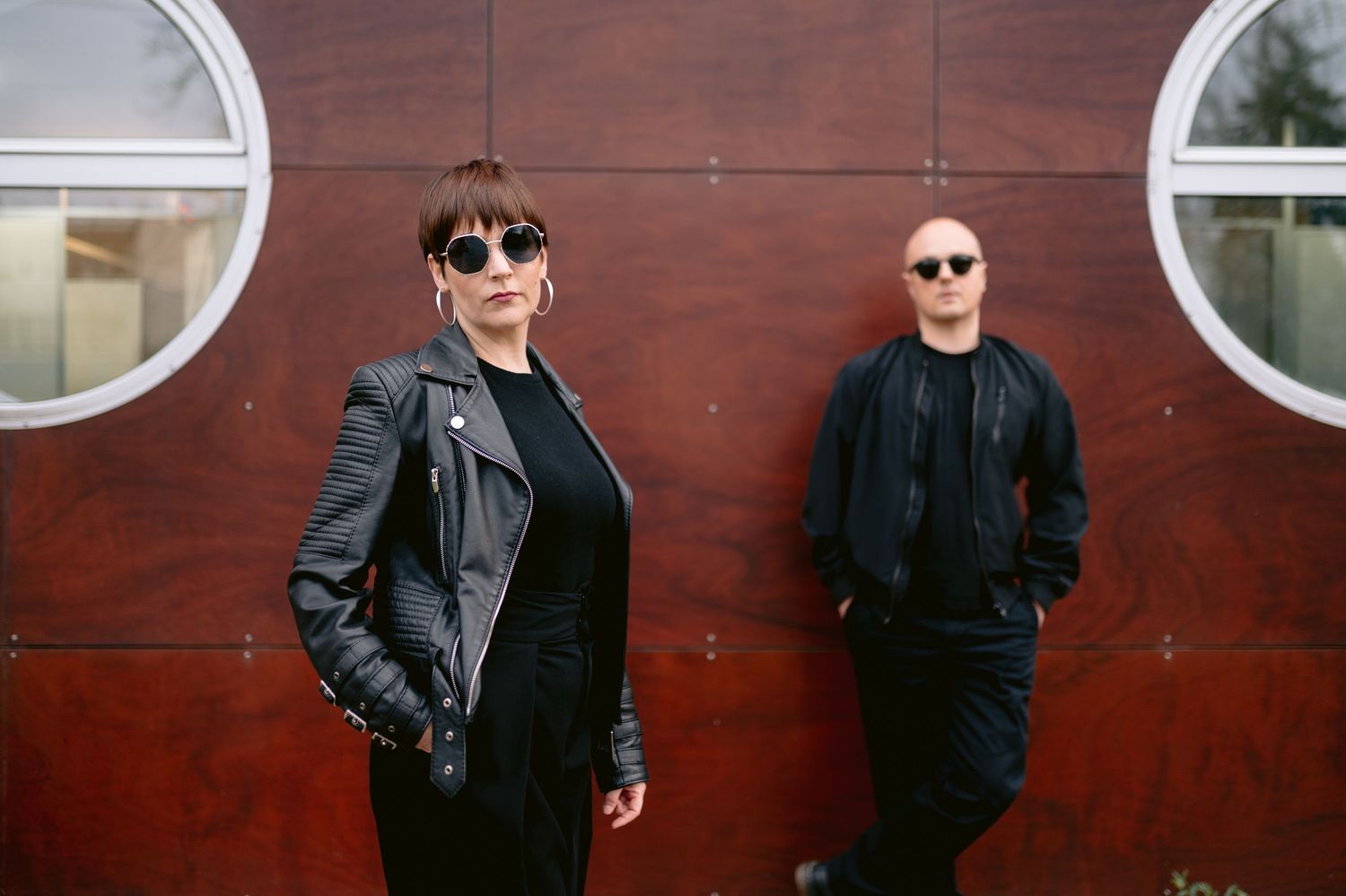 Two musicians in black outfits pose against a dark red metal wall with circular windows.