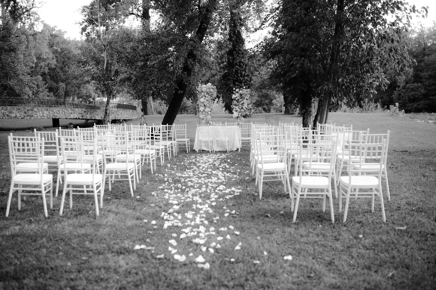 Black and white outdoor wedding ceremony setup with white chairs and rose petal aisle under lush trees.
