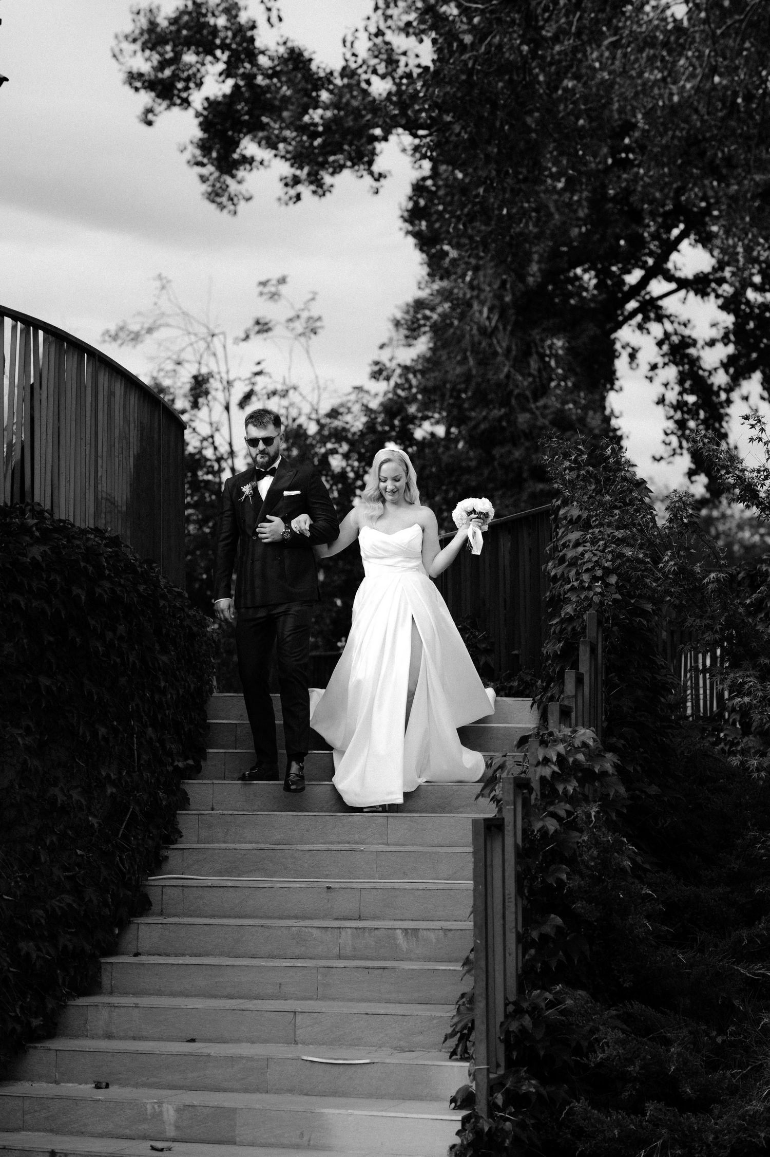 Black and white photo of a bride and groom walking down outdoor steps, bride holding bouquet, surrounded by lush greenery.