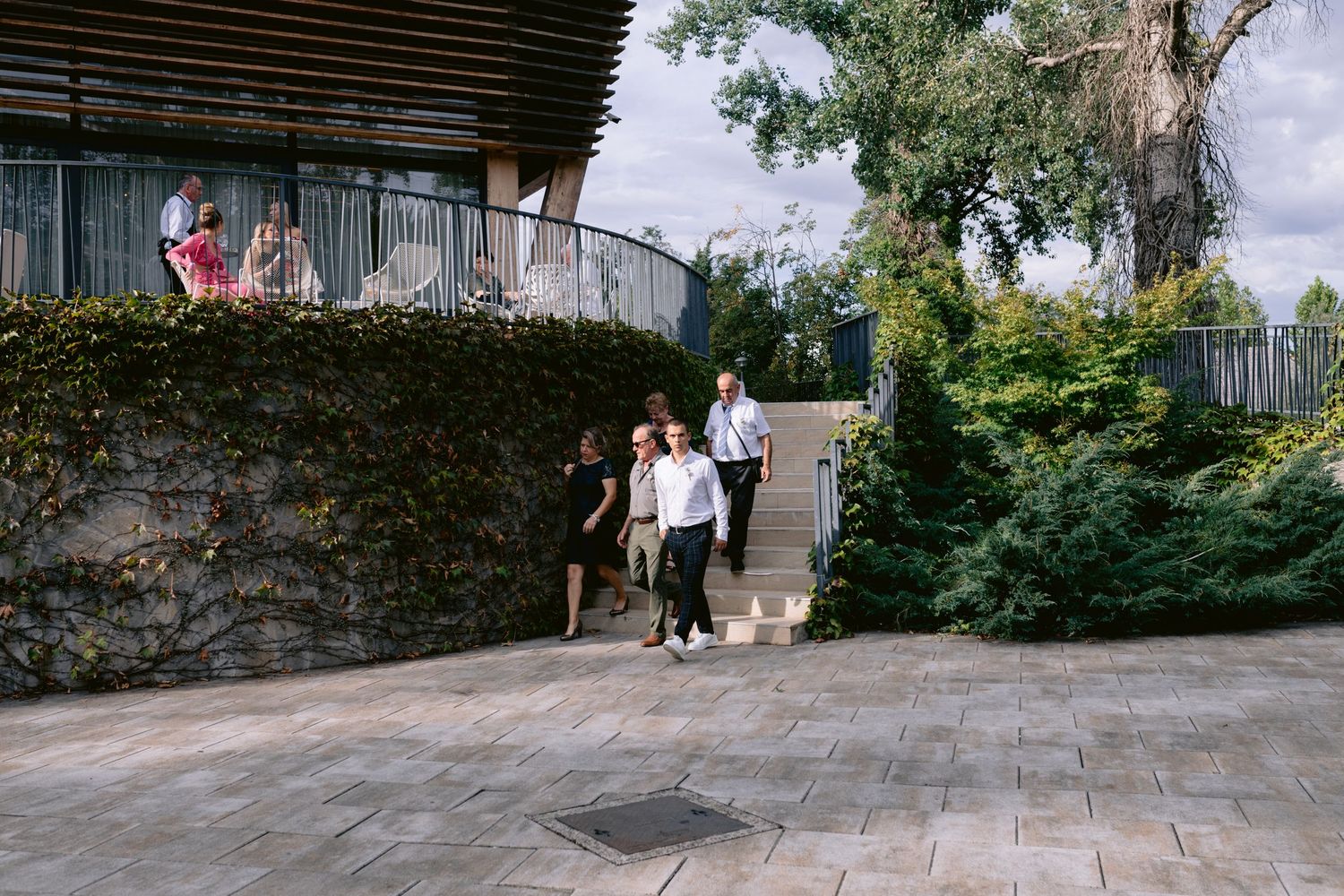 People walking on a stone patio near an ivy-covered curved building surrounded by lush green trees.