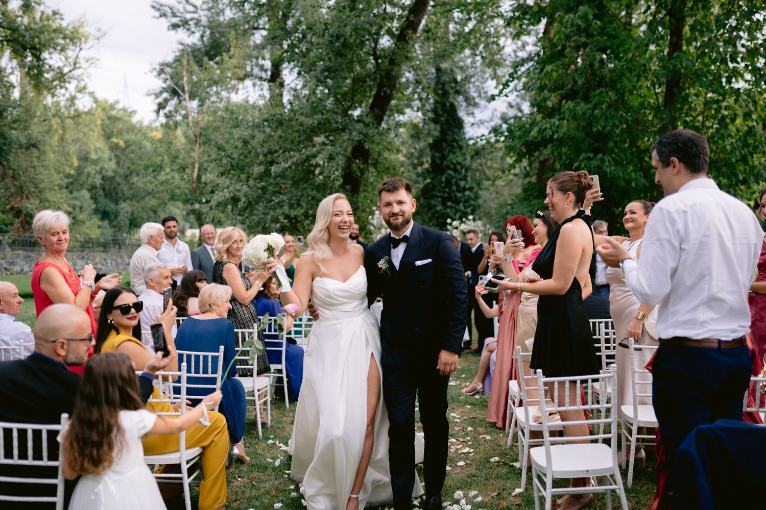 Bride and groom walk down the aisle at an outdoor wedding ceremony surrounded by cheering guests and lush green trees.