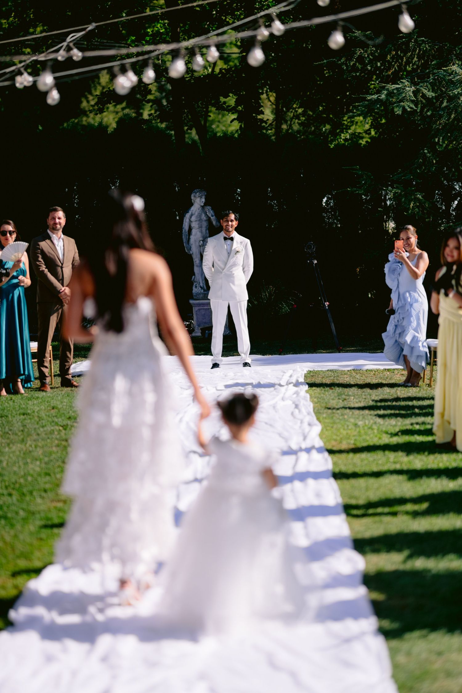 Bride in white gown walks down outdoor aisle with flower girl as groom waits at altar during garden wedding ceremony.