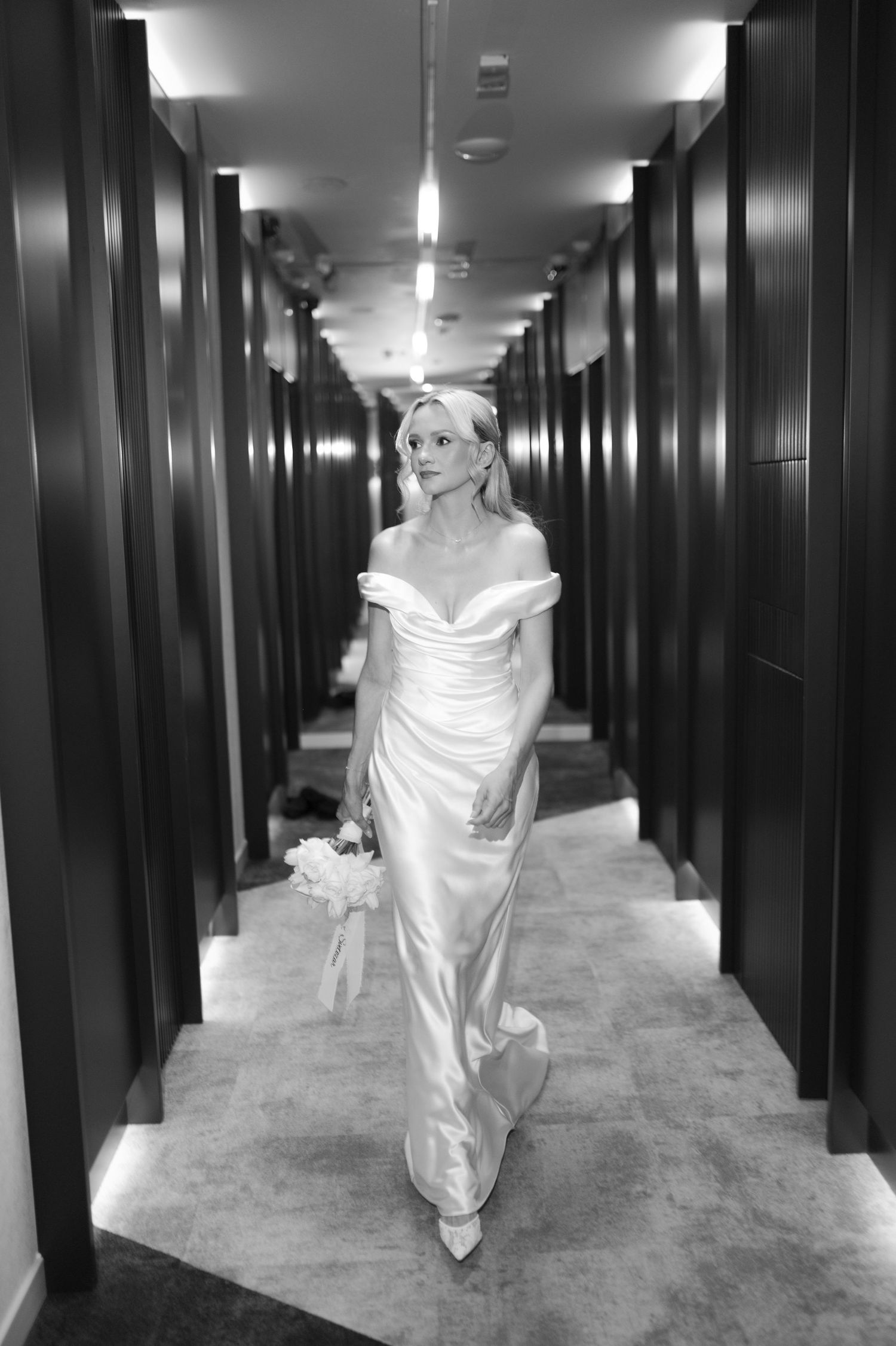 Black and white photo of a bride in an off-shoulder satin gown walking down a long hallway holding a bouquet.