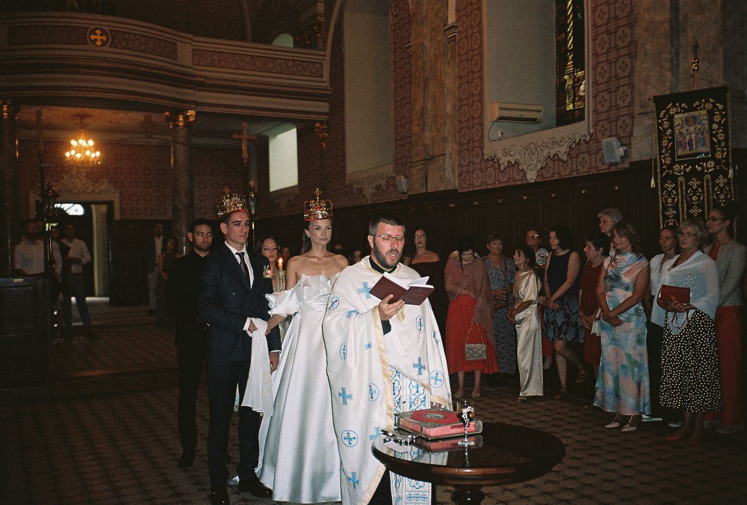 Wedding party members in light blue dresses and white suits walk in a processional line through an ornate venue.