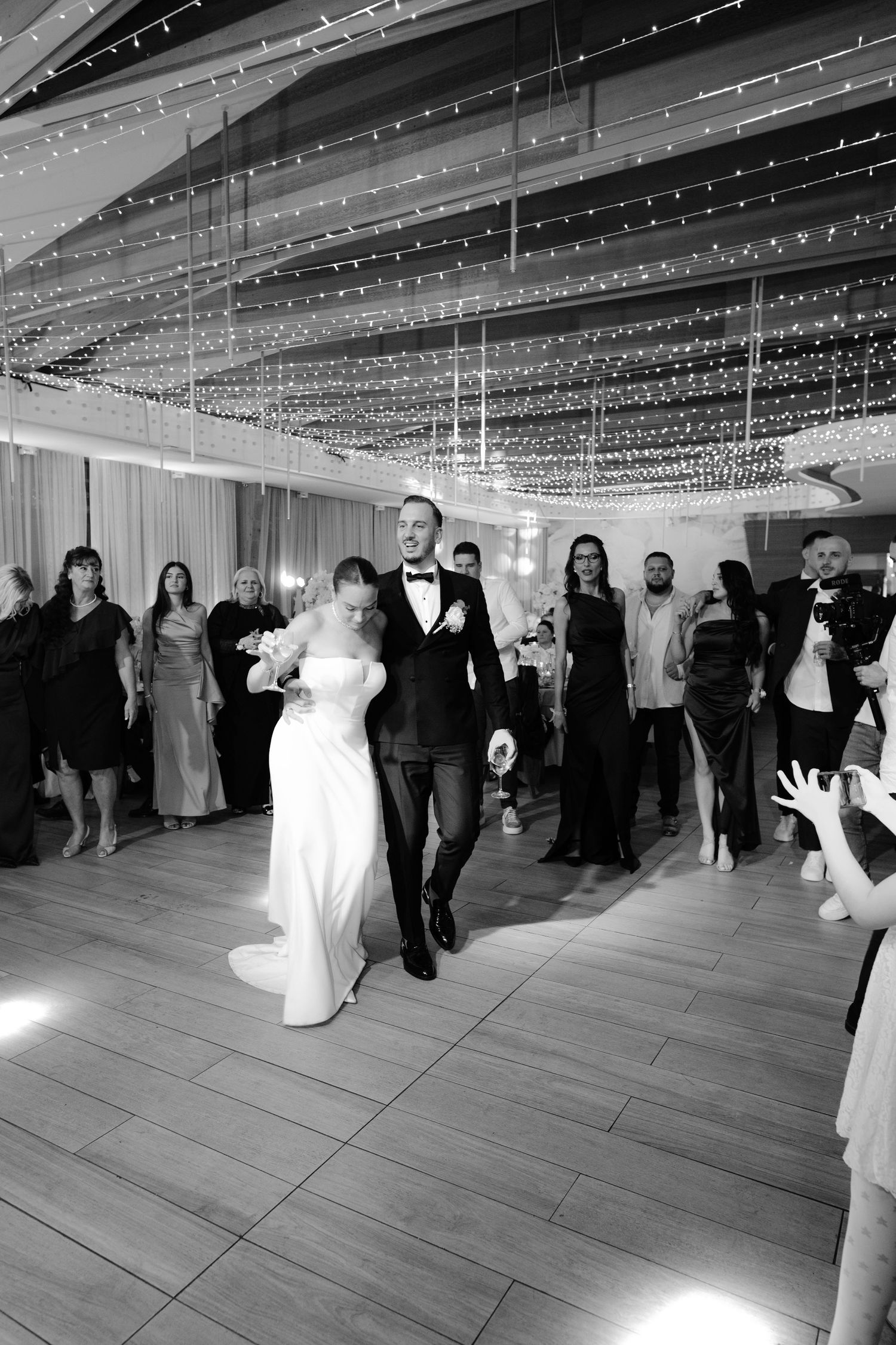 Wedding guests dance under twinkling string lights on a spacious dance floor at an elegant reception venue.