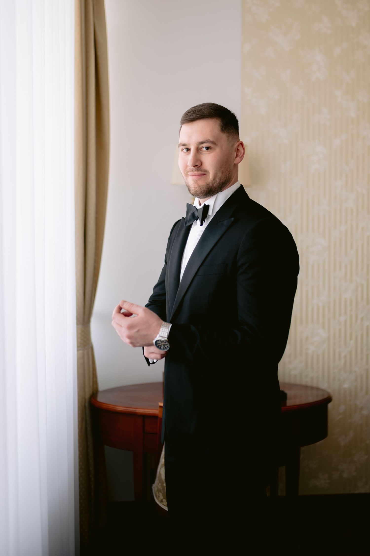 Man in black suit and tie checks his phone while standing in a room with beige wallpaper and curtains.