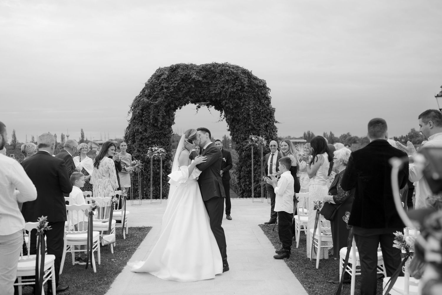 Black and white photo of a wedding ceremony under a floral arch with guests seated on either side of the aisle.
