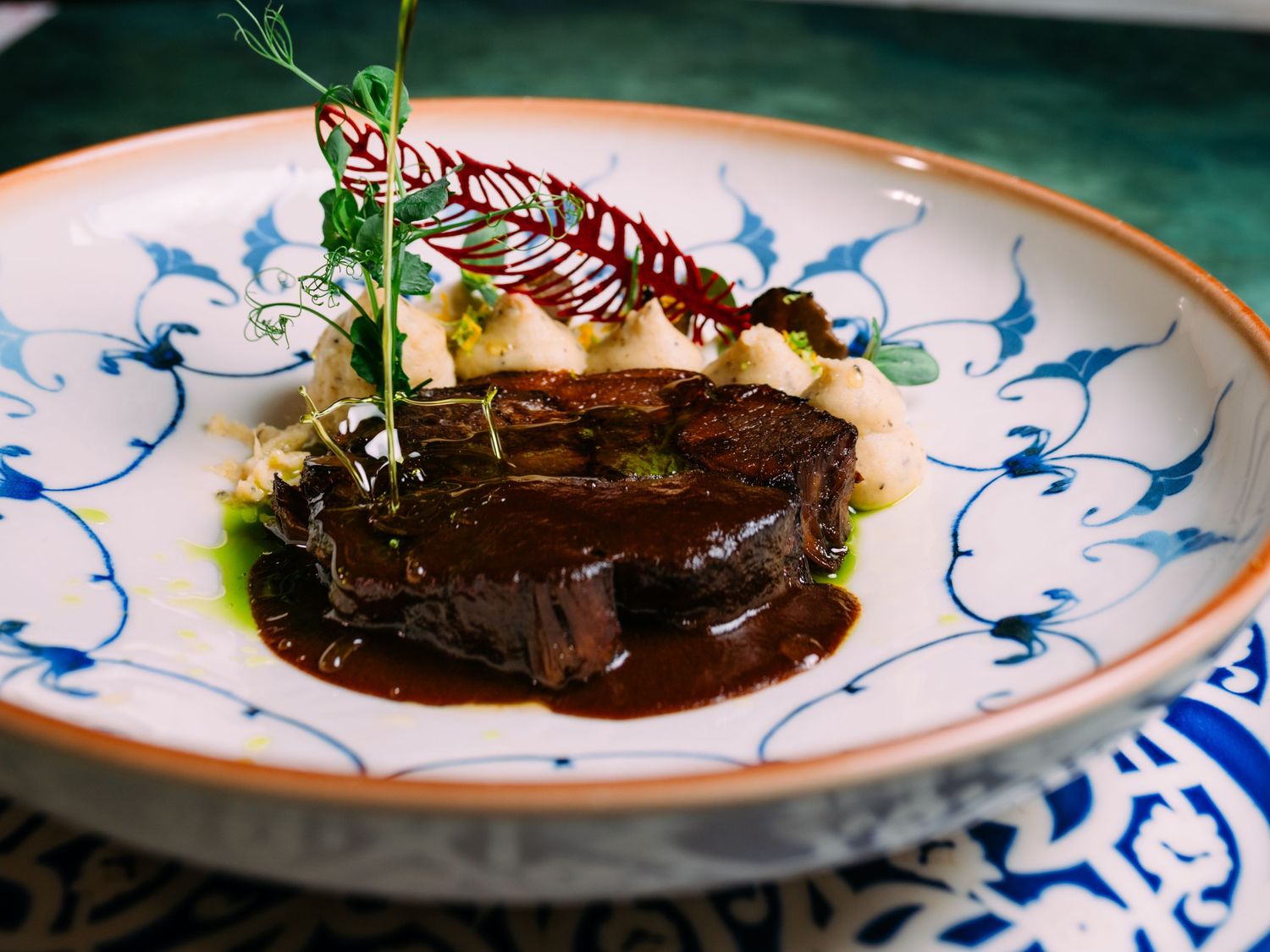 Plated braised beef short ribs with dark rich sauce and fresh herbs served on white plates with blue designs.