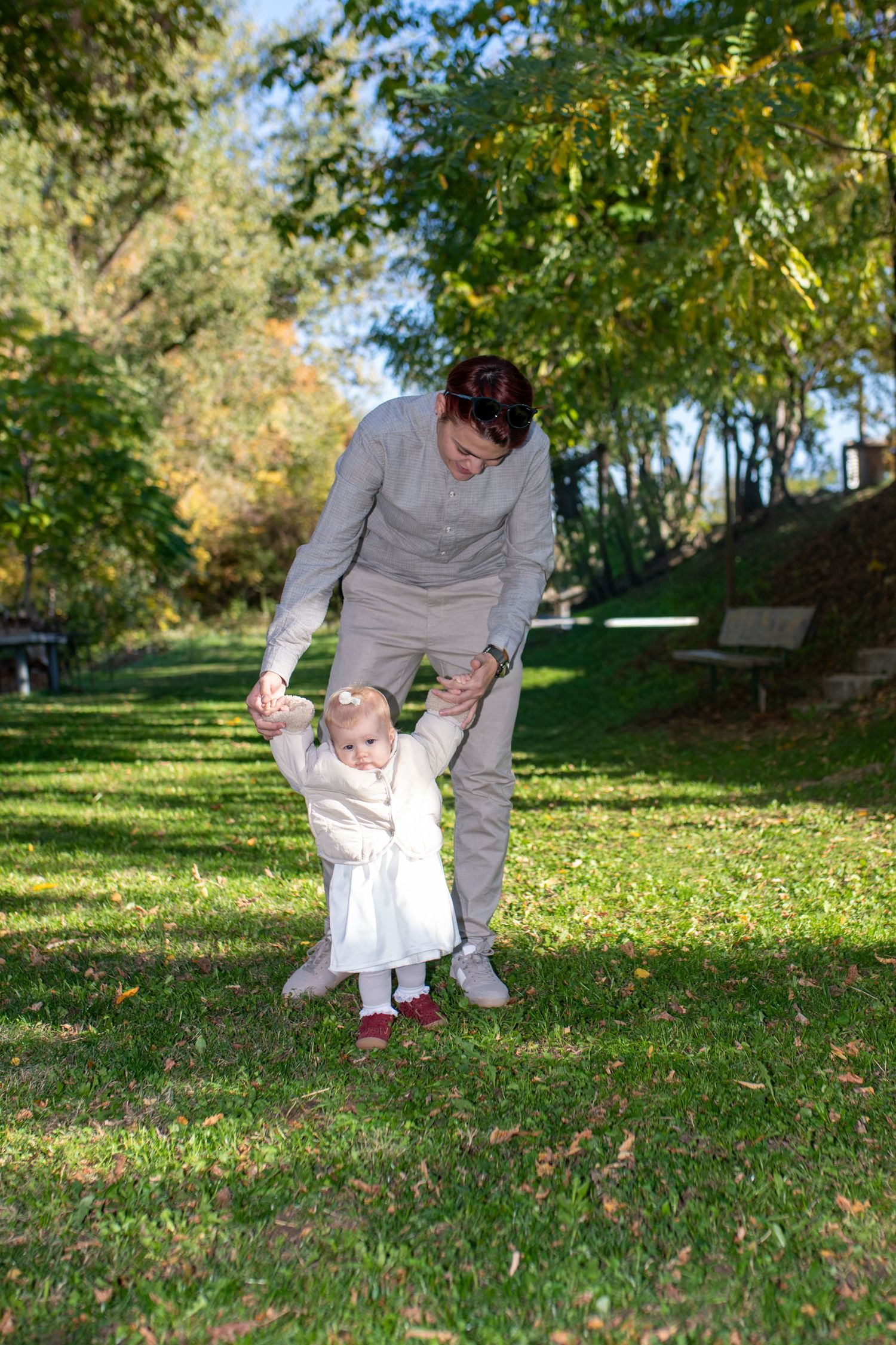 A parent holds the hands of a toddler in a white dress taking first steps on a sunny grassy park lawn surrounded by autumn trees.