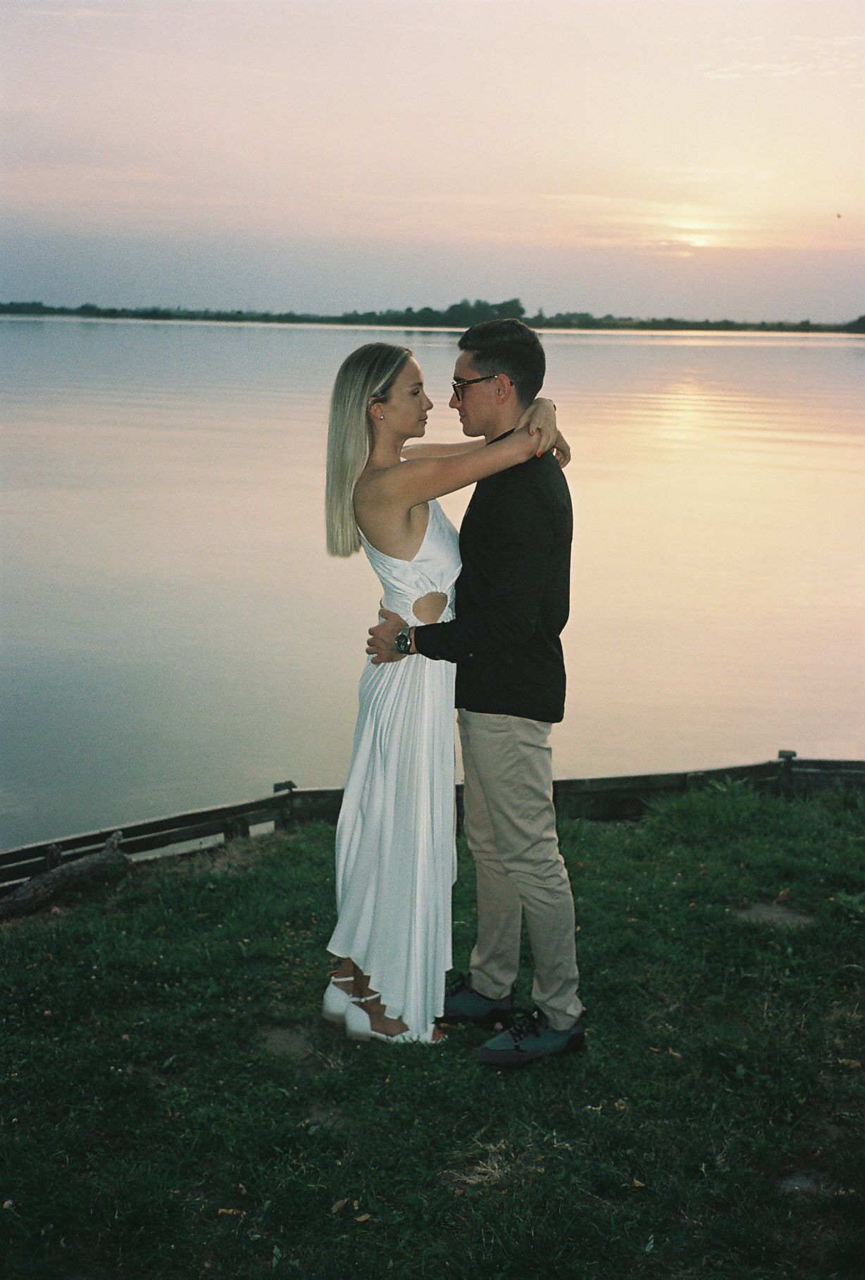 A couple shares a romantic moment at sunset by a serene lake, with the woman wearing a white dress and man in khakis.