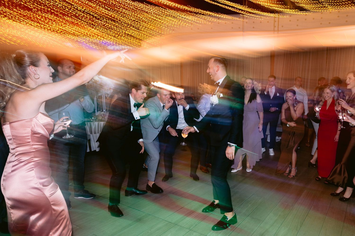 Wedding reception guests dance together on a wooden floor under warm glowing lights during an evening celebration.