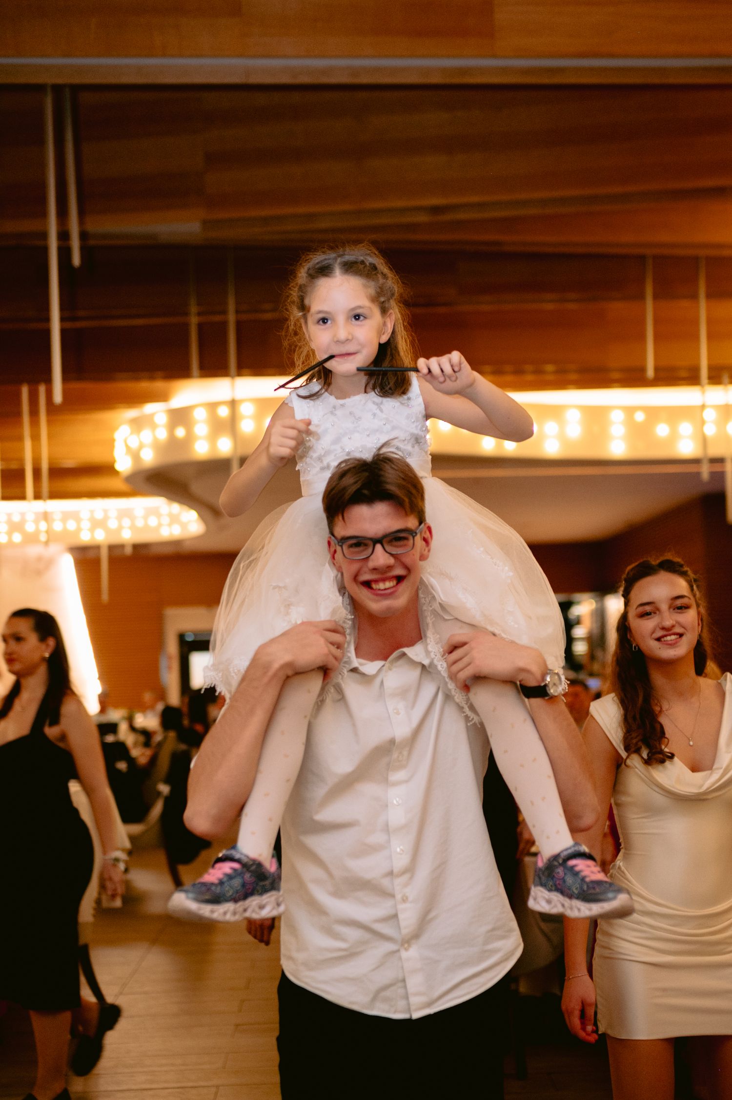 A smiling teen boy carries a young girl on his shoulders at a festive indoor event, with a girl in white dress behind them.
