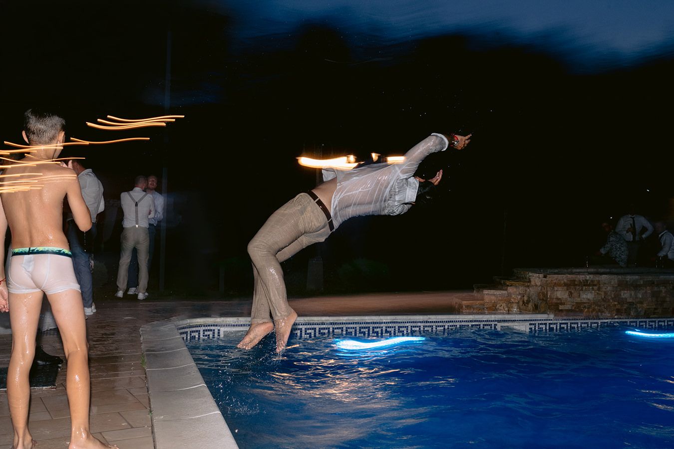 Man diving into illuminated pool at night party while guests watch nearby.