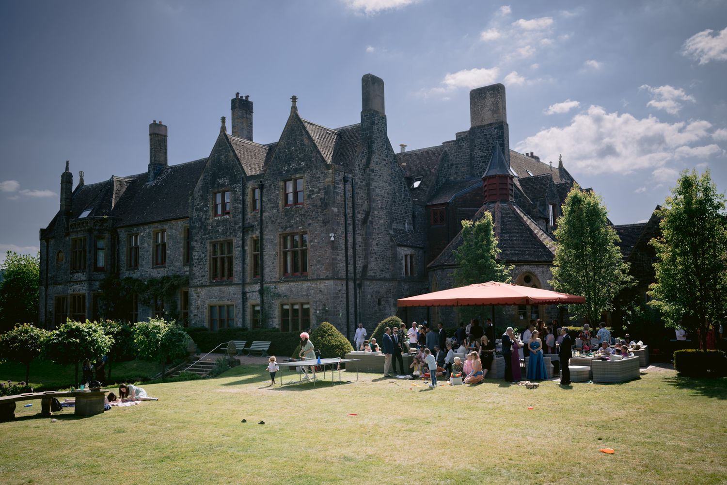 Elegant stone manor house wedding venue with guests gathered on sunny lawn under red parasols.