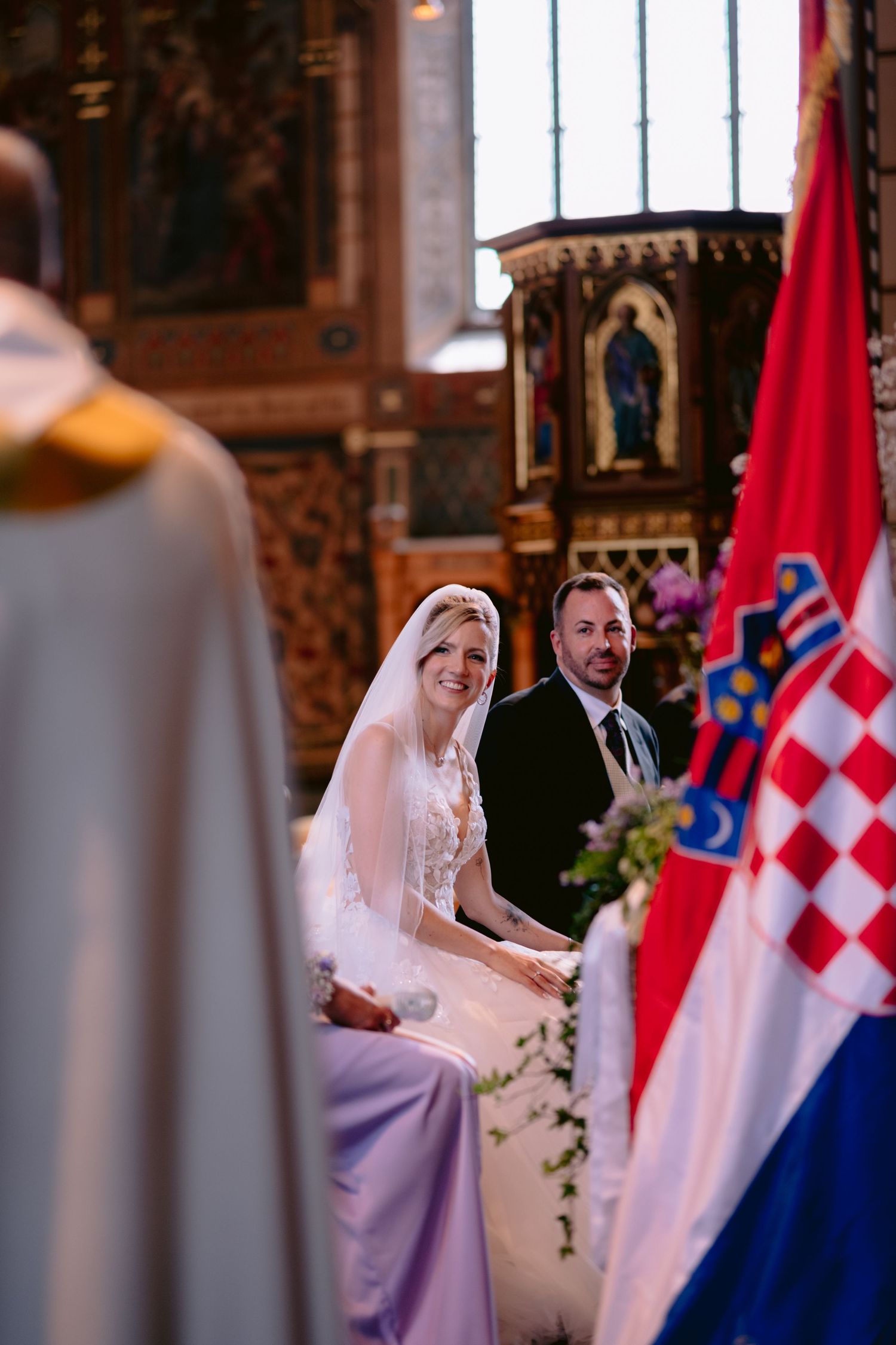 Bride and groom seated at church altar during wedding ceremony with Croatian flag and ornate interior.