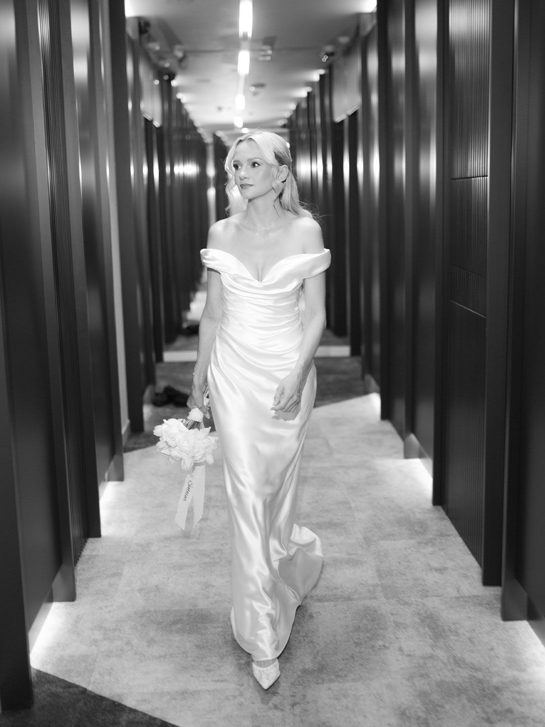 Elegant bride in off-shoulder white satin gown walks corridor holding bouquet, black and white photo.