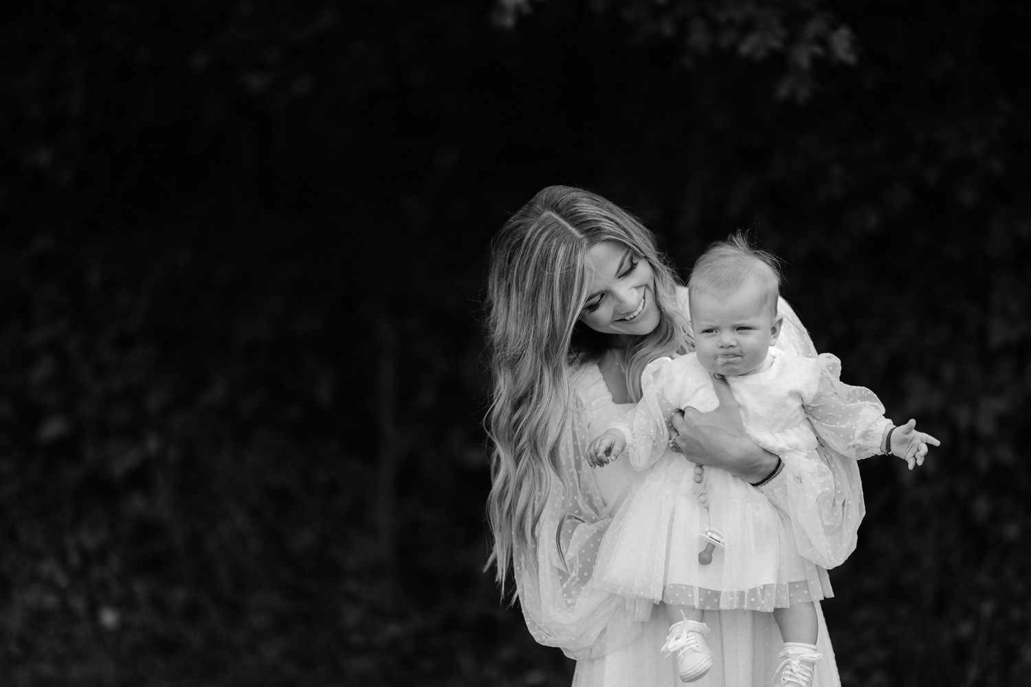 Black and white photo of a blonde woman in white dress tenderly holding a baby in a lush garden setting.