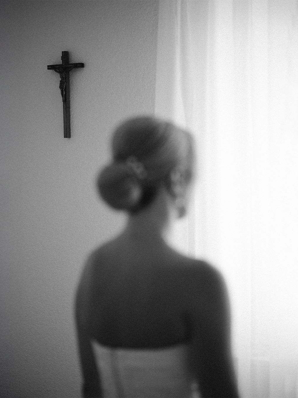 Blurred black and white photo of a bride in a strapless gown facing a crucifix on a wall near sheer curtains.
