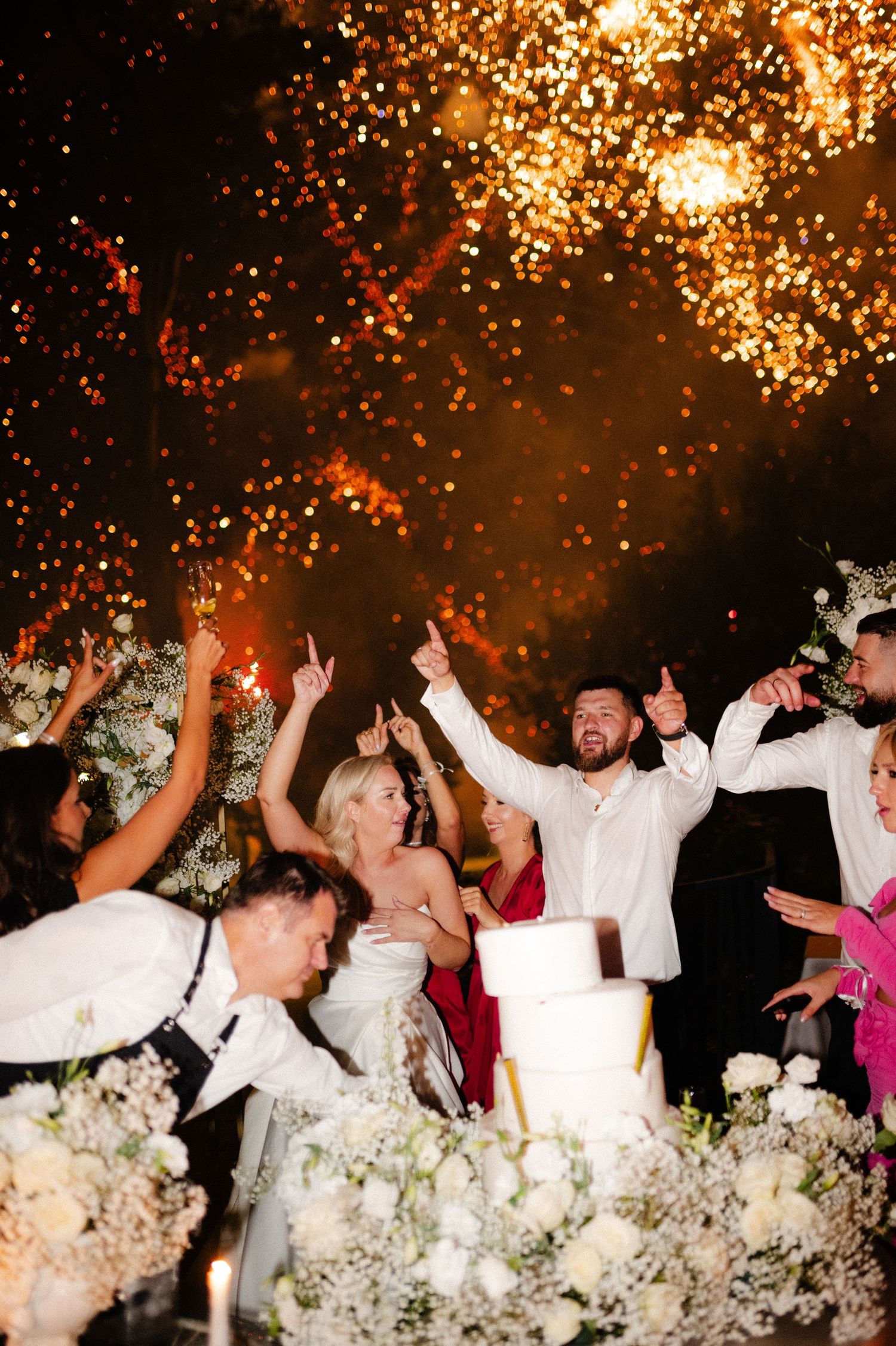 Bride and groom celebrate with guests amid fireworks and white floral arrangements at an elegant outdoor wedding reception.