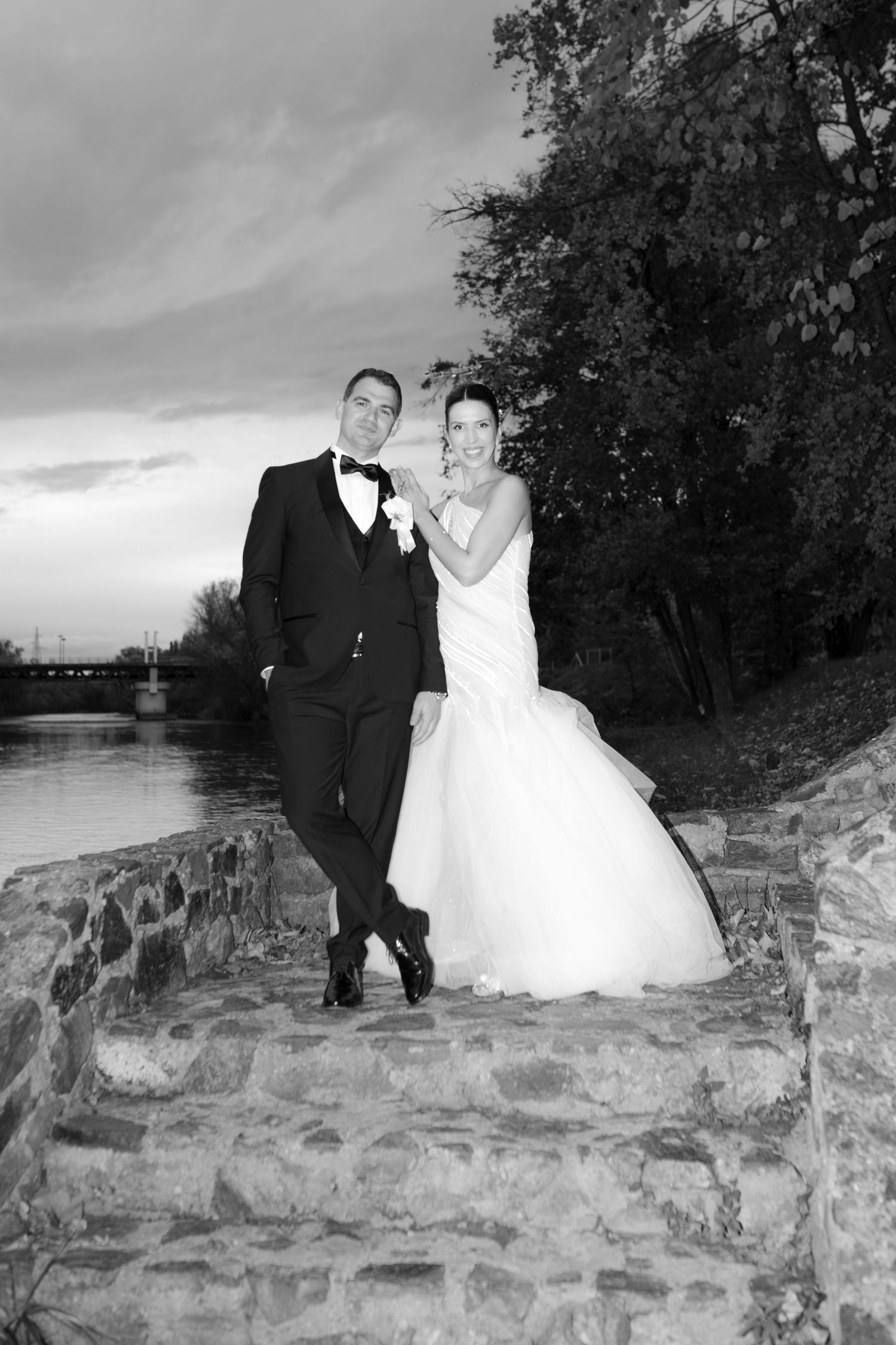Black and white portrait of newlywed couple in formal attire posing by riverside stone steps at dusk.