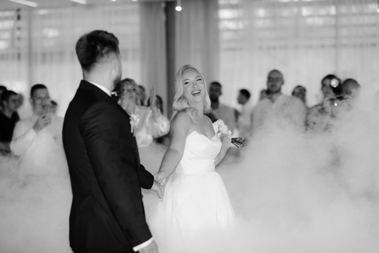 Black and white photo of bride and groom sharing first dance surrounded by fog machine smoke at elegant wedding reception.
