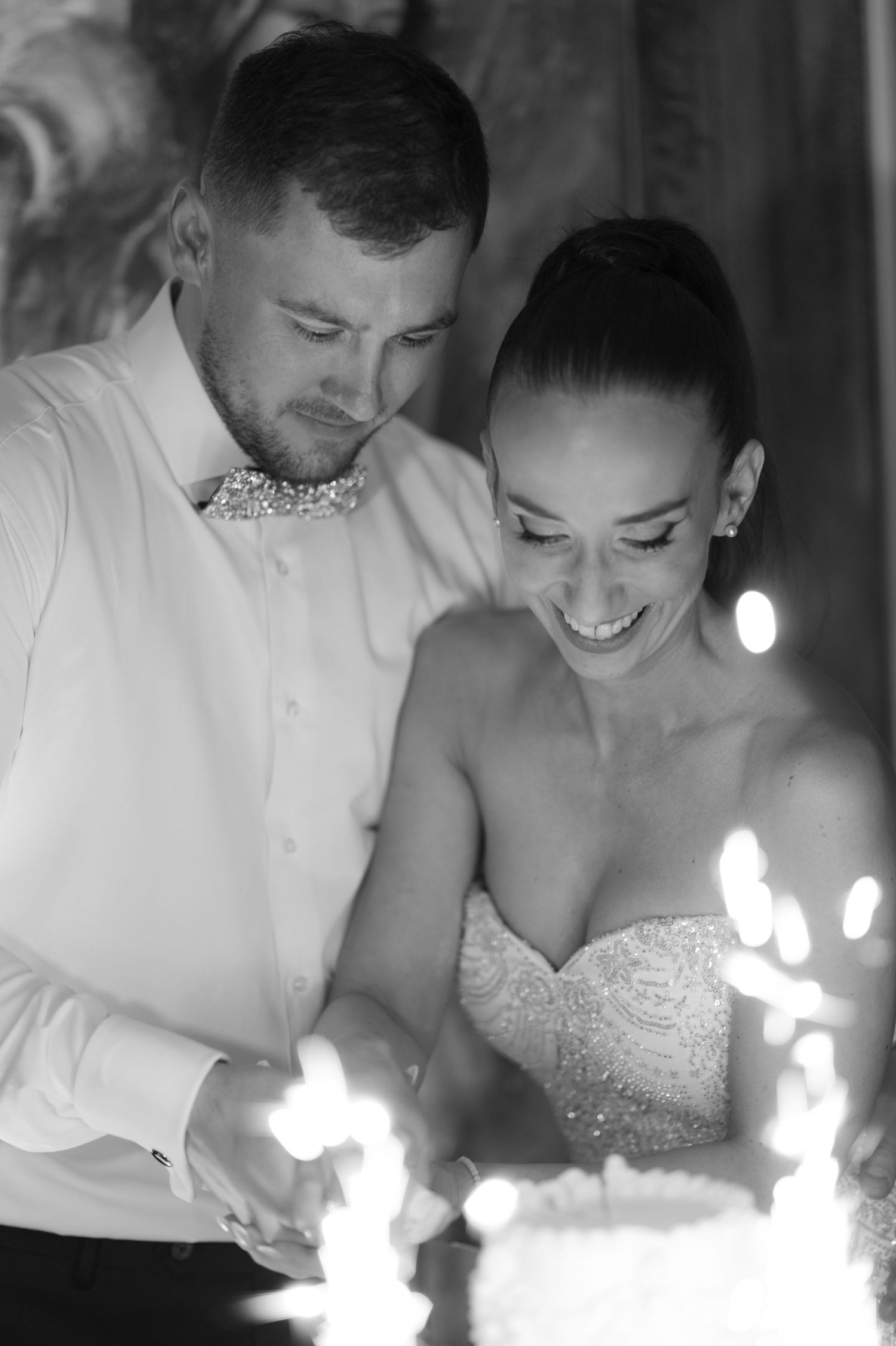 A bride and groom share a joyful moment while cutting their wedding cake illuminated by sparklers in black and white.