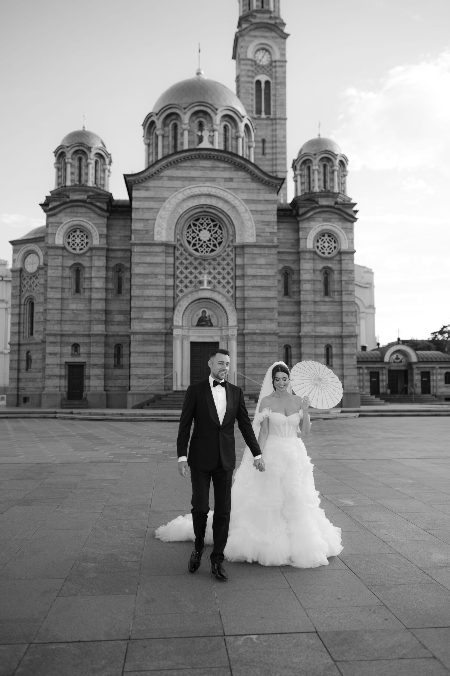 Black and white wedding photo of bride and groom holding hands before grand orthodox church.