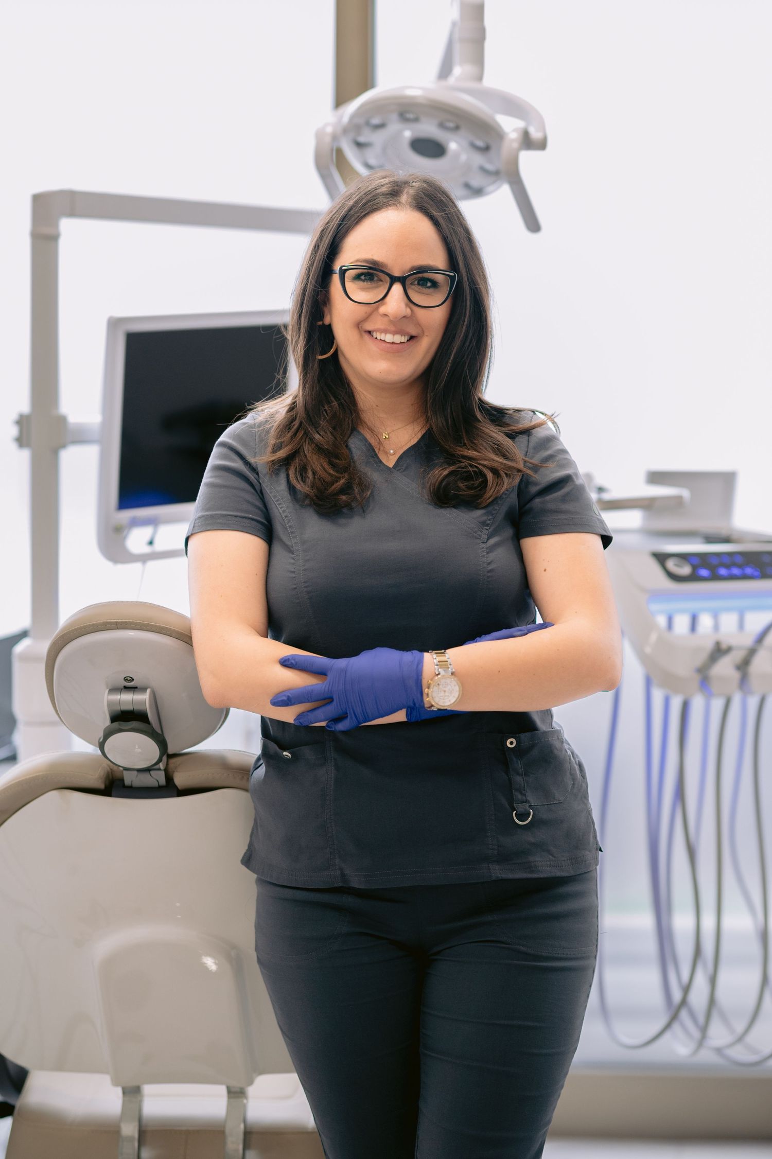 Female dentist in dark scrubs and blue gloves smiling in a modern dental office with equipment visible behind her.