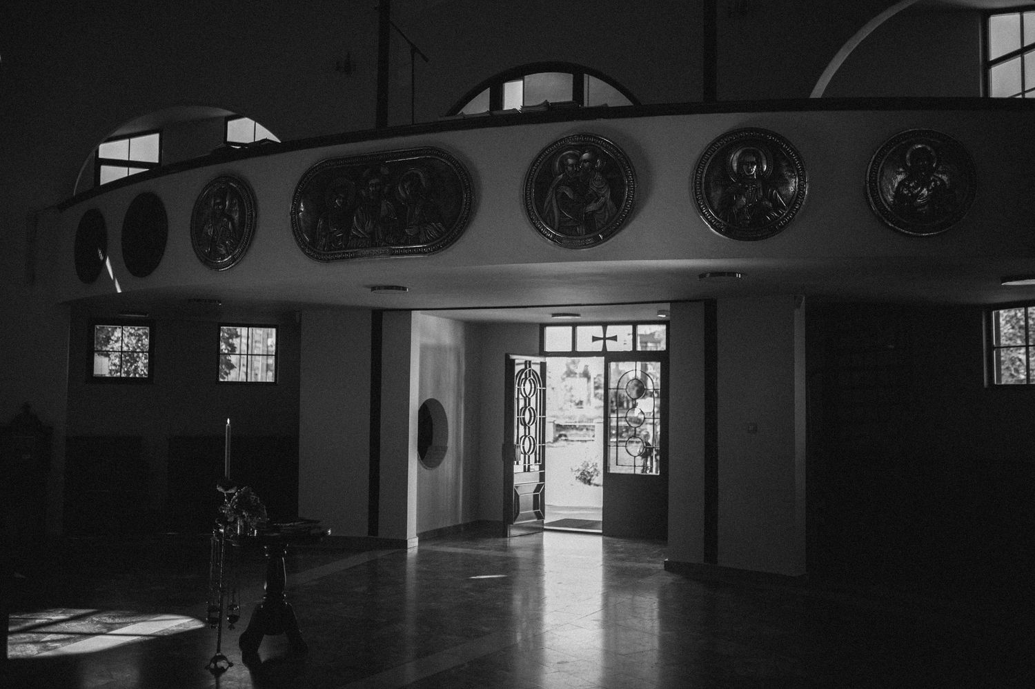 Sunlight streams through an open doorway into a dim church interior with round icon medallions mounted along a curved balcony.