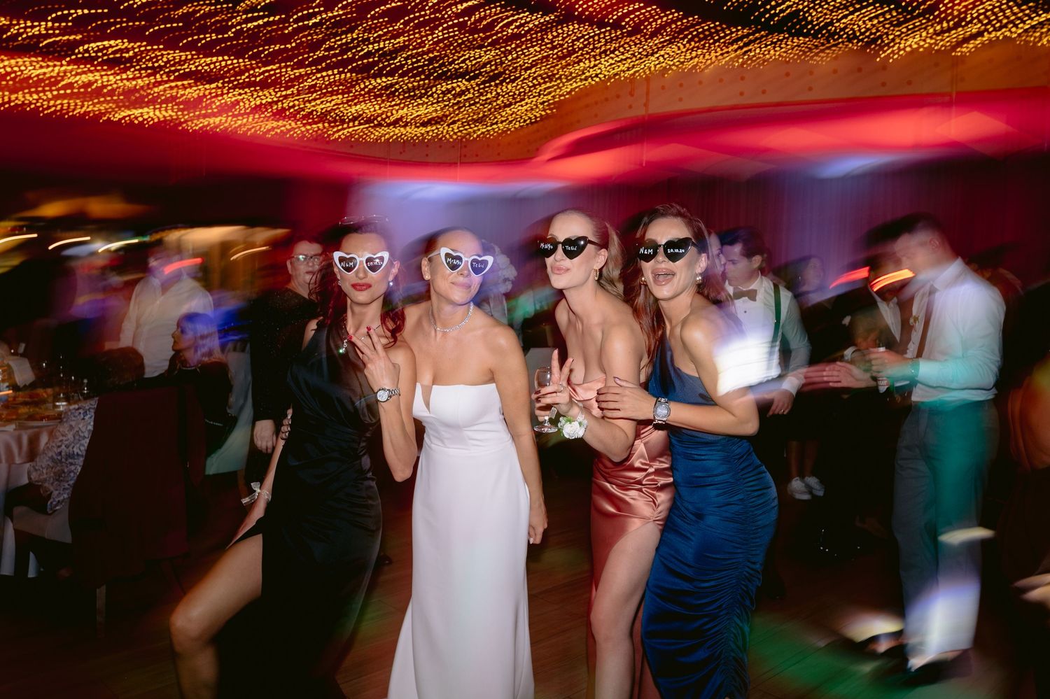 Four women in elegant dresses and sunglasses dancing at a lively wedding reception with colorful lights.
