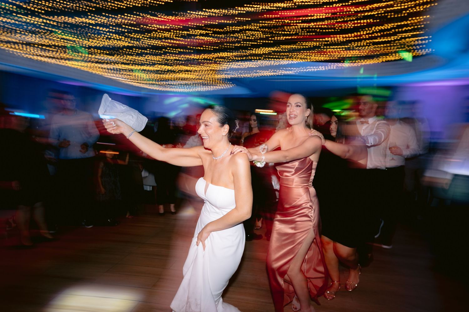 Motion-blurred dancers twirl on a dance floor under colorful string lights creating a festive atmosphere at an evening event.