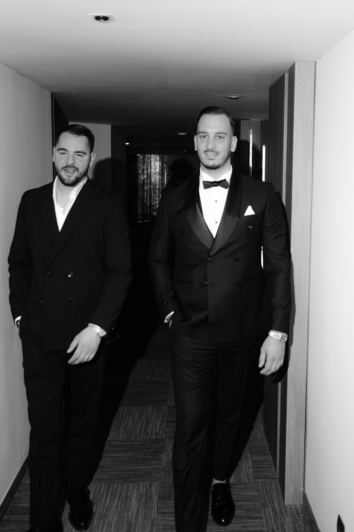 Groomsmen in black tuxedos and bow ties walk down a hallway before a formal event.
