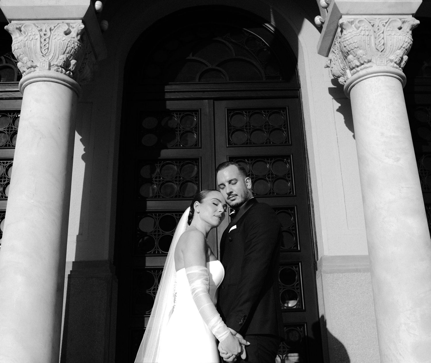 Black and white photograph of a couple in formal attire posing between elegant white columns.