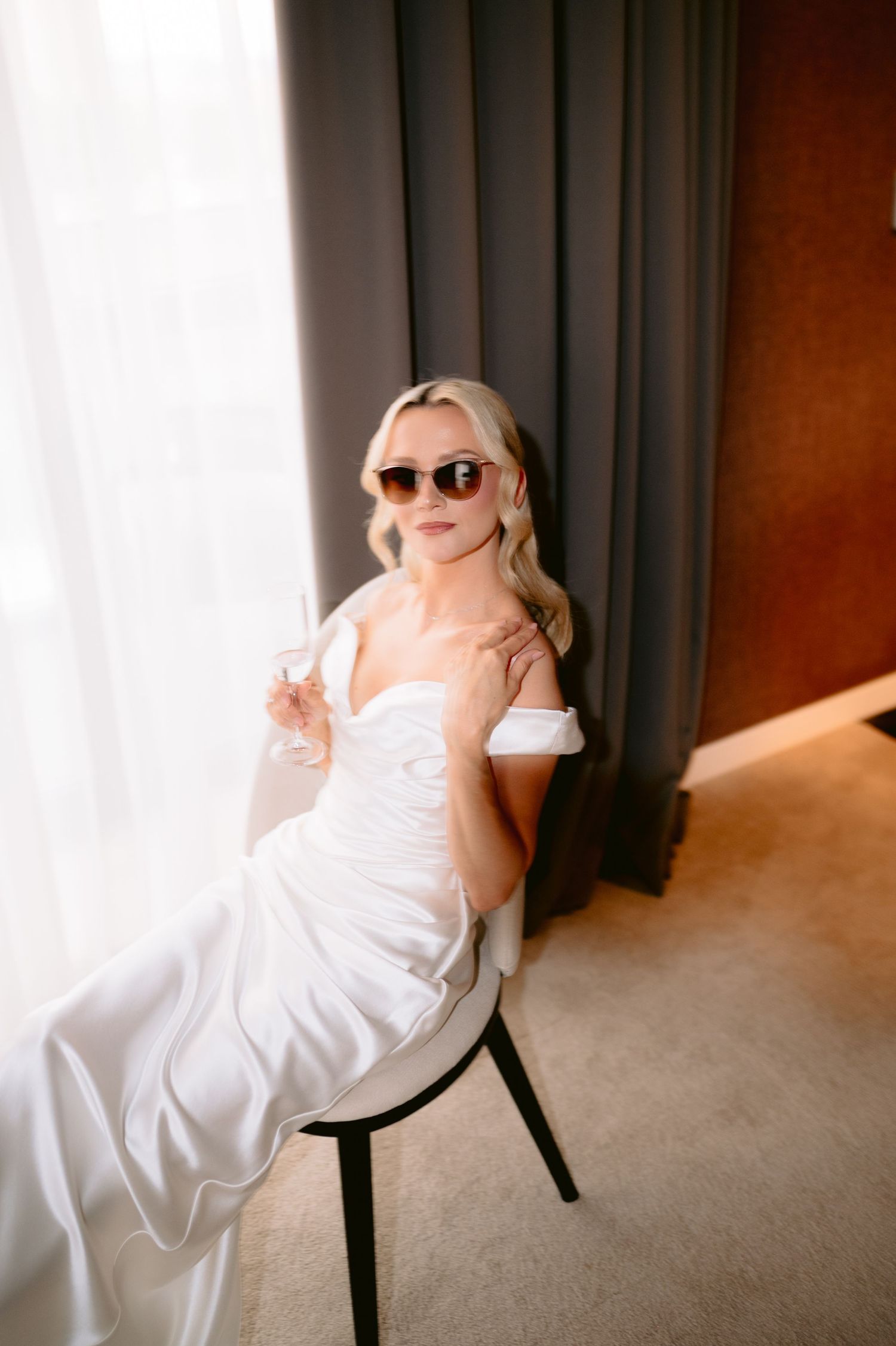 Blonde bride in white off-shoulder gown and sunglasses seated elegantly by a window with gray curtains.