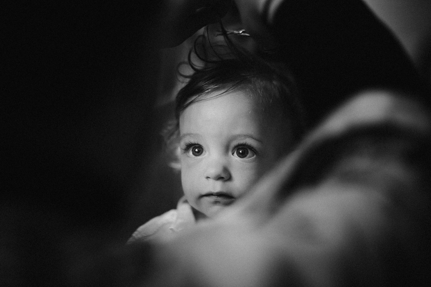 Black and white portrait of a curious baby with dark hair gazing forward, captured in soft focus lighting.