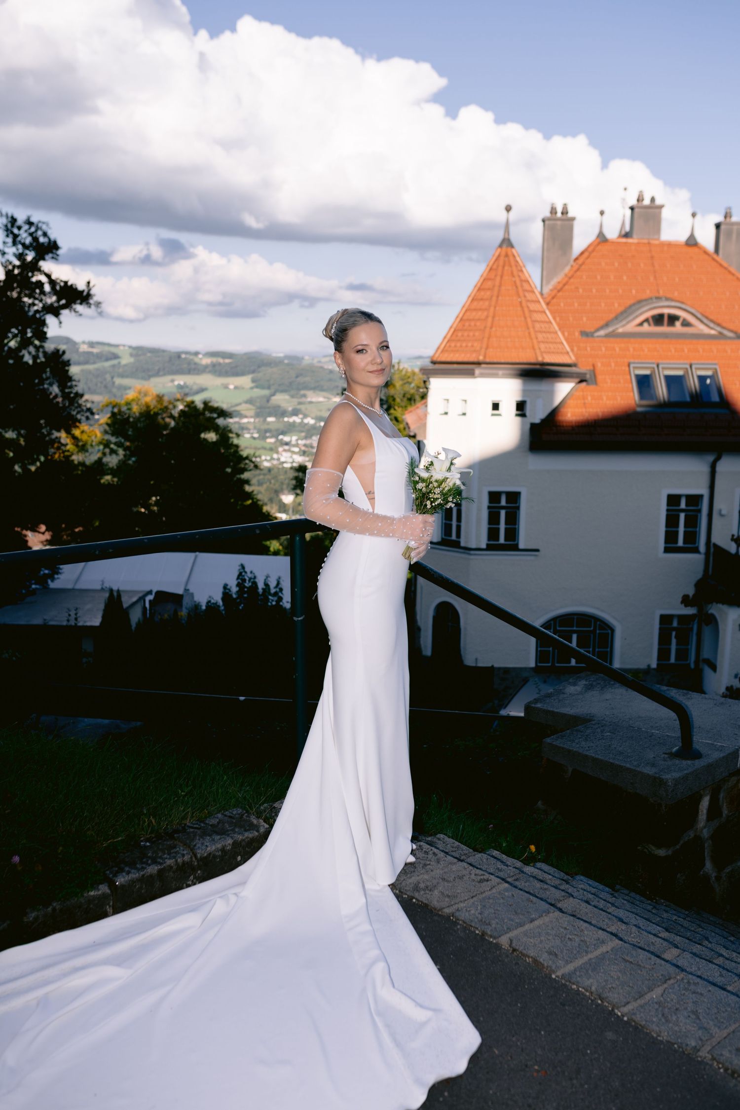Bride in elegant white gown with train poses on terrace of European castle with orange rooftops and scenic hills.