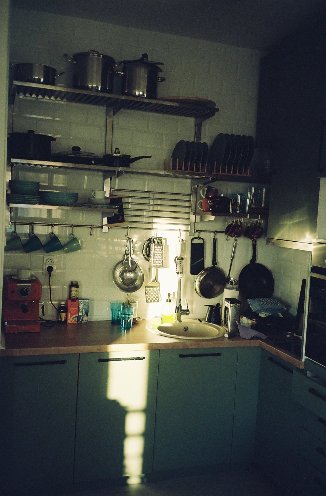 Dimly lit kitchen corner with hanging pots and pans on wall-mounted shelves and sunlight streaming through window.