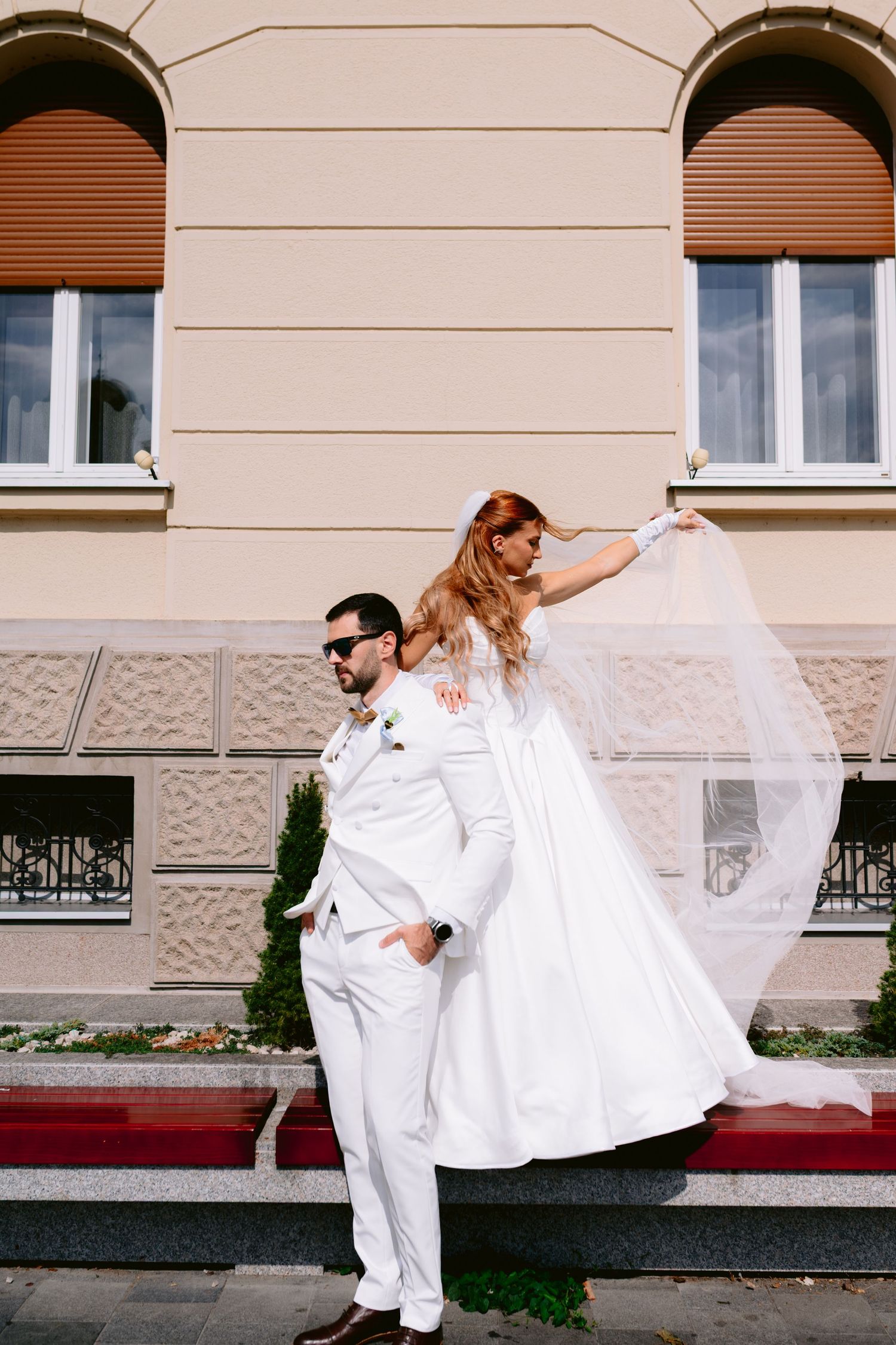 Mladoženja u bijelom odijelu nosi mladu u vjenčanici s dugim velom ispred elegantne zgrade tokom sunčanog dana.