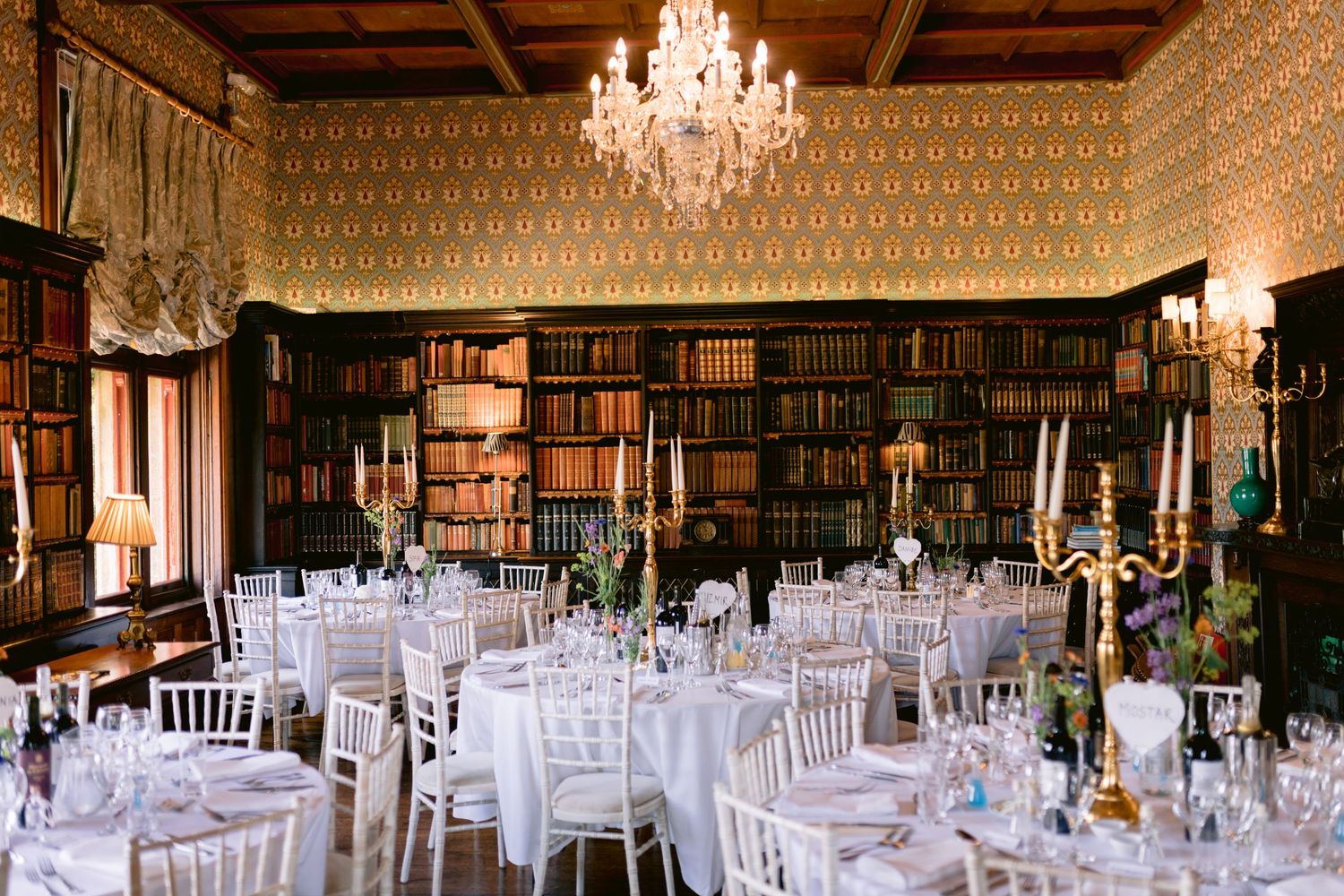 Elegant library wedding reception with chandeliers and white table settings surrounded by bookshelves.