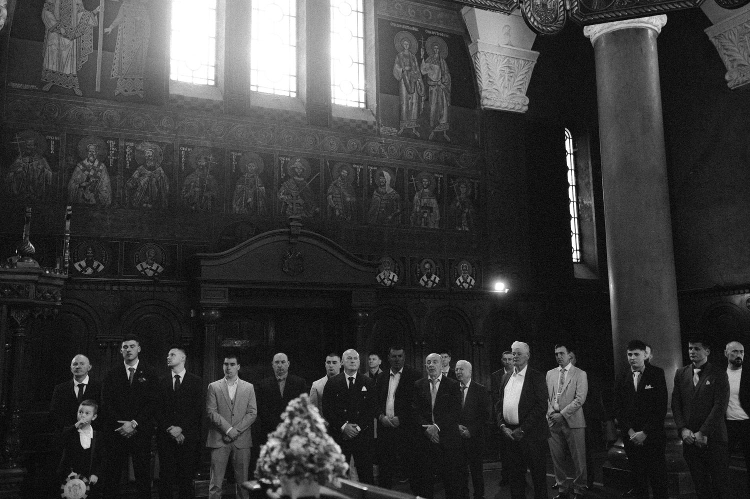 A black and white photograph shows a formal gathering in a grand hall with ornate walls and columns illuminated by window light.
