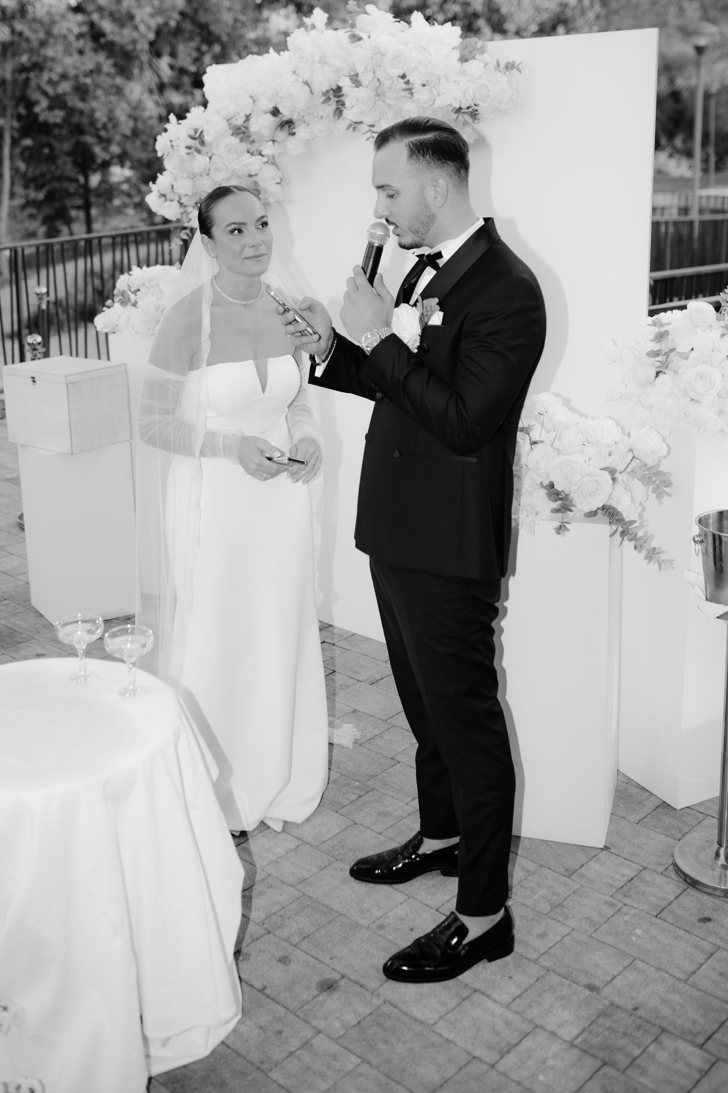 A black and white wedding photo of a couple sharing a moment during their reception under a floral arch.