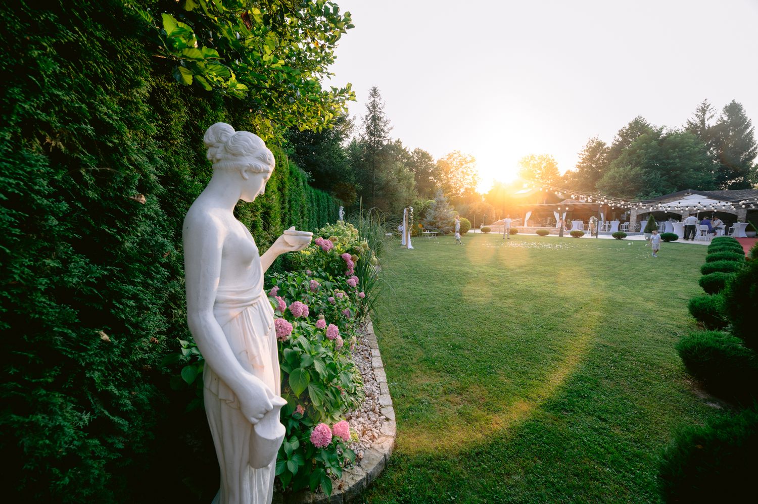 Garden statue in white dress against sunset with floral landscaping.