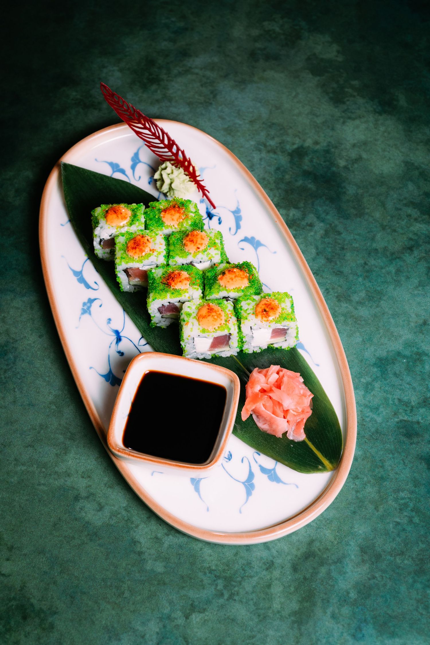 Overhead view of green tobiko sushi rolls on a banana leaf with soy sauce and pickled ginger on an oval ceramic plate.