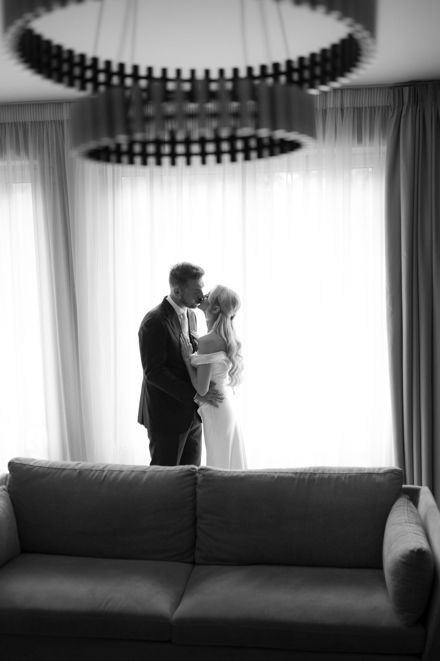 Black and white photo of a couple embracing and kissing in an elegant room with a modern chandelier and sofa.