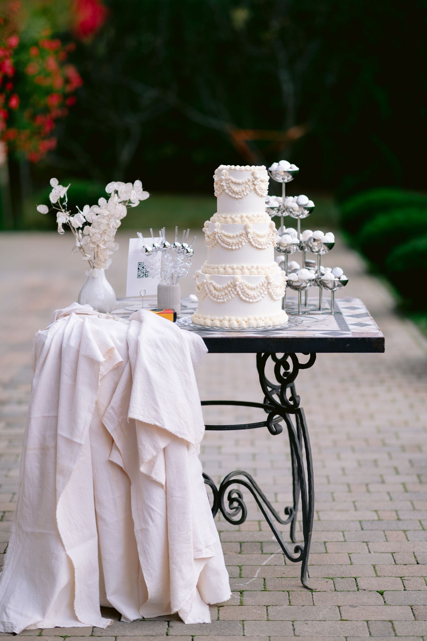 Elegant white wedding cake and decor displayed on wrought iron table with flowing white tablecloth on brick patio.
